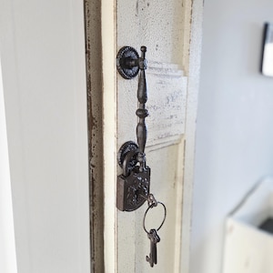 May include: Ornate, dark metal door handle and lock with keys attached. The handle is vertically oriented and attached to a distressed, off-white door. The lock and keys have a vintage, antique appearance.