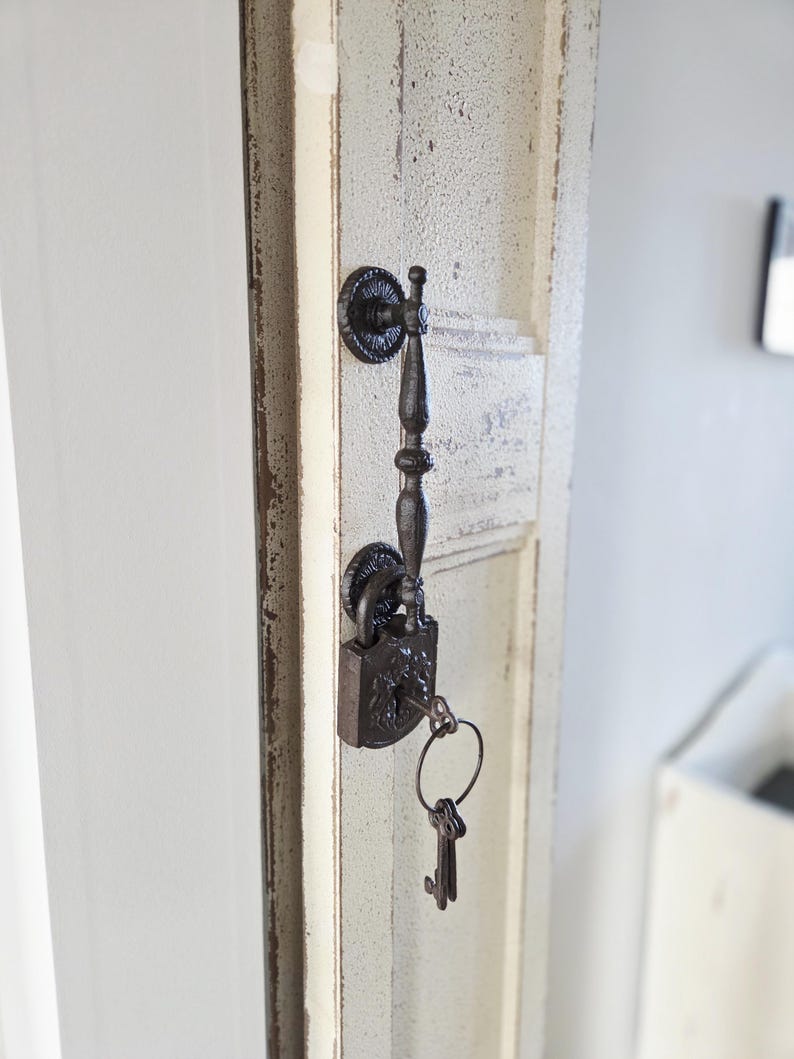 May include: Ornate, dark metal door handle and lock with keys attached. The handle is vertically oriented and attached to a distressed, off-white door. The lock and keys have a vintage, antique appearance.
