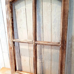 May include: A rustic wooden window frame with a distressed finish. The frame is divided into four panes and has an arched top. The wood has a warm, brown tone with white accents, creating a vintage aesthetic. The window frame is displayed against a light-colored wooden background.