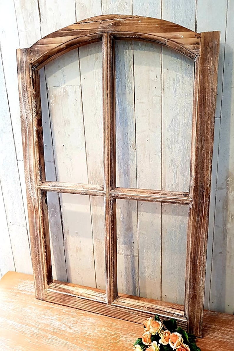 May include: A rustic wooden window frame with a distressed finish. The frame is divided into four panes and has an arched top. The wood has a warm, brown tone with white accents, creating a vintage aesthetic. The window frame is displayed against a light-colored wooden background.