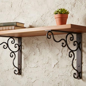 Op de afbeelding: Een houten wandplank ondersteund door twee zwarte metalen beugels met sierlijk krulwerk. De plank bevat twee gestapelde boeken met bruine ruggen en een kleine groene vetplant in een terracotta-kleurige pot, gemonteerd aan een muur.