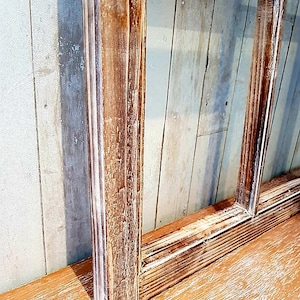 May include: A rustic, rectangular wooden frame with a distressed finish. The frame is made of light brown wood with visible grain and white paint accents. The frame is set against a light blue and white wooden background.