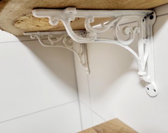 May include: A white metal shelf bracket featuring an ornate scrollwork design with curved detailing. The bracket is mounted to a wall and supports a thick, natural-edged wooden shelf board, showcasing a sturdy construction and a matte finish.