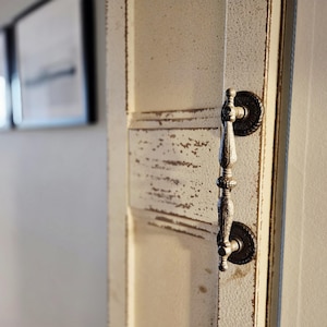 May include: Close-up of a distressed white door with a decorative, ornate metal handle. The door has a weathered, vintage appearance with visible paint chipping. The handle is dark metal with intricate detailing, adding a touch of elegance to the rustic design.