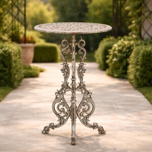 May include: Ornate, antique-style outdoor side table. The round tabletop features a detailed floral pattern. The table has a distressed white finish, with intricate scrollwork on the legs and support. The table is approximately 28 inches tall.