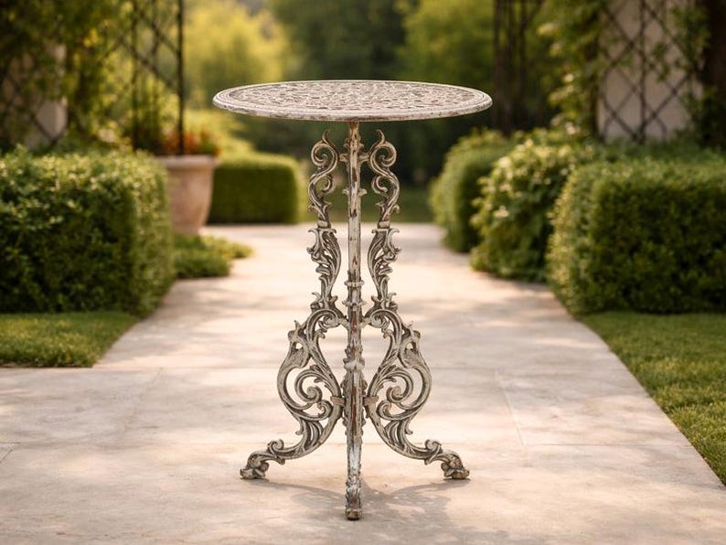 May include: Ornate, antique-style outdoor side table. The round tabletop features a detailed floral pattern. The table has a distressed white finish, with intricate scrollwork on the legs and support. The table is approximately 28 inches tall.