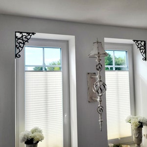 May include: Two black metal corner brackets with intricate scrollwork designs mounted in the upper corners of two white window frames. The brackets feature a decorative, curved pattern that extends from the corner along the wall and window edge.