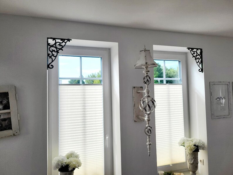 May include: Two black metal corner brackets with intricate scrollwork designs mounted in the upper corners of two white window frames. The brackets feature a decorative, curved pattern that extends from the corner along the wall and window edge.