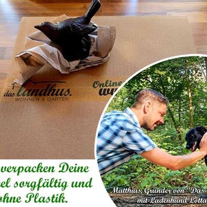 May include: A black cast iron bird figurine resting on crumpled brown paper atop a cardboard box labelled das landhus WOHNEN & GARTEN. A circular inset shows a person in a blue plaid shirt petting a black dog in a forest with text below.