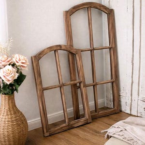 Può includere: Due cornici di finestre ad arco in legno con finitura rustica sono appoggiate a un muro. Le cornici hanno un motivo a griglia e sono in legno marrone. Un vaso intrecciato con fiori rosa è nelle vicinanze.
