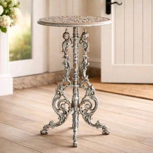 Puede incluir: Mesa auxiliar ornamental de tres patas con acabado blanquecino envejecido. La mesa redonda presenta un patrón decorativo. La mesa es de metal y es adecuada para uso en interiores o exteriores.