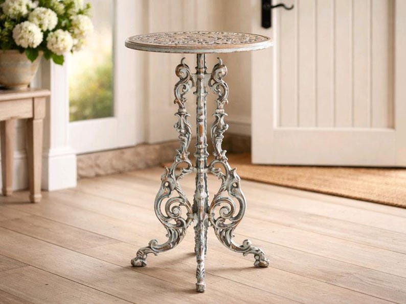 May include: Ornate, three-legged side table with a distressed, off-white finish. The round tabletop features a decorative pattern. The table is made of metal and is suitable for indoor or outdoor use.