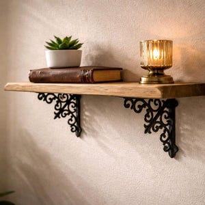 Op de afbeelding: Een houten plank met een bruin, ledergebonden boek, een kleine potplant en een glazen kandelaar. De plank wordt ondersteund door twee zwarte, sierlijke metalen beugels, waardoor een rustieke en decoratieve presentatie ontstaat.