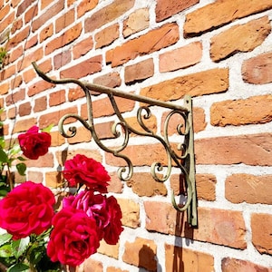 Op de afbeelding: Een sierlijke, antieke metalen plantenhouder is bevestigd aan een verweerde bakstenen muur. De houder heeft decoratief krulwerk en een haak om een mand of plantenbak op te hangen. Felrode rozen bloeien in de buurt.