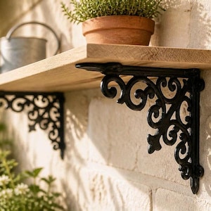 Può includere: Una mensola in legno chiaro sostenuta da due staffe in metallo nero con intricati motivi a volute. La mensola è montata su una parete in pietra strutturata e sostiene un vaso in terracotta con una pianta verde e un annaffiatoio in metallo.