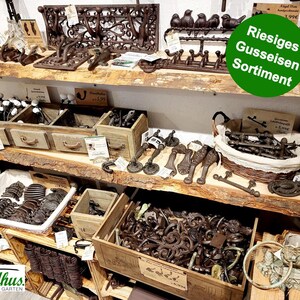 May include: A display of cast iron home decor items, including hooks, drawer pulls, and decorative accents. The items are in various shades of brown and are arranged on wooden shelves. The image includes the text "Riesiges Gusseisen Sortiment".