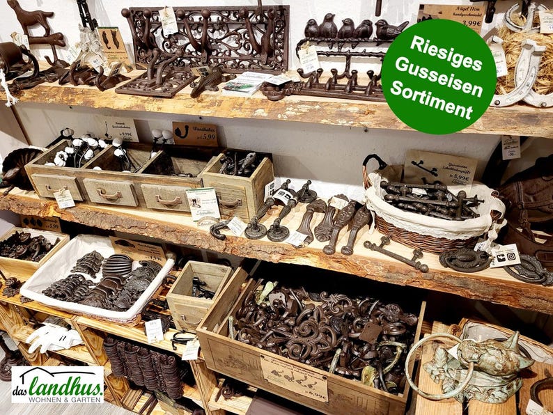 May include: A display of cast iron home decor items, including hooks, drawer pulls, and decorative accents. The items are in various shades of brown and are arranged on wooden shelves. The image includes the text "Riesiges Gusseisen Sortiment".