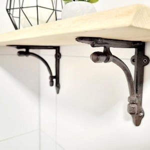 Peut inclure: Une étagère en bois clair soutenue par deux supports en métal foncé et texturé. Les supports présentent un design incurvé avec des boutons décoratifs et des trous de fixation. L'étagère est fixée contre un mur blanc, avec un objet en fil métallique.