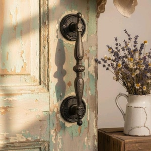 May include: Ornate black metal door handle with a decorative design, mounted on a weathered, peeling paint door. The door's paint is a mix of blue, green, and beige. A white pitcher with flowers sits on a wooden crate.