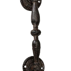 May include: A rustic, cast iron door pull with a dark brown finish. The handle has an ornate, vertical design with decorative circular backplates. The hardware is likely for a cabinet or door.
