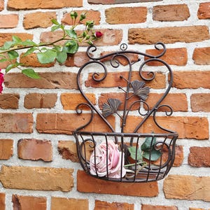 Puede incluir: Una cesta de pared decorativa de hierro forjado con diseño de corazón, llena de una rosa rosa y vegetación, cuelga de una pared de ladrillo. La cesta presenta trabajos decorativos en metal y es un acento encantador para arreglos florales.