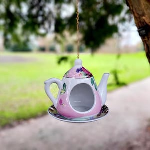 Könnte beinhalten: Ein hängendes Vogelhäuschen aus Keramik in Form einer Teekanne mit rosa Körper und Blumenmuster, weißem Henkel und Ausguss sowie einer runden Öffnung an der Vorderseite. Die Teekanne steht auf einer passenden Untertasse und hängt an einer dünnen goldfarbenen Kette.