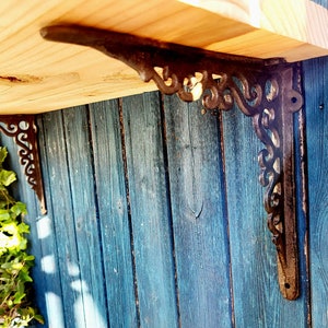 Könnte beinhalten: Zwei verzierte schwarze Metall-Regalbretter mit einem dekorativen Schriftrollenmuster sind an einer blauen Holzwand montiert. Die Halterungen tragen ein Holzregal.