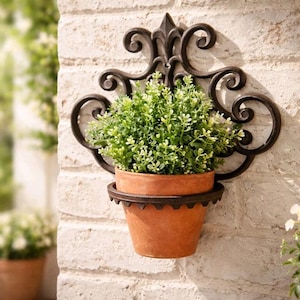 Op de afbeelding: Een decoratieve wandplanter met een terracotta pot gevuld met groen gebladerte. De pot wordt vastgehouden door een donkerbruine, sierlijke metalen beugel tegen een witte bakstenen muur. De beugel heeft een krullend ontwerp.