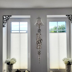 May include: A vertical wall-mounted decorative ornament featuring a distressed white finish with dark accents. The piece consists of a small lampshade-style top, two spherical cage-like sections, and a pointed finial base, positioned between two windows.
