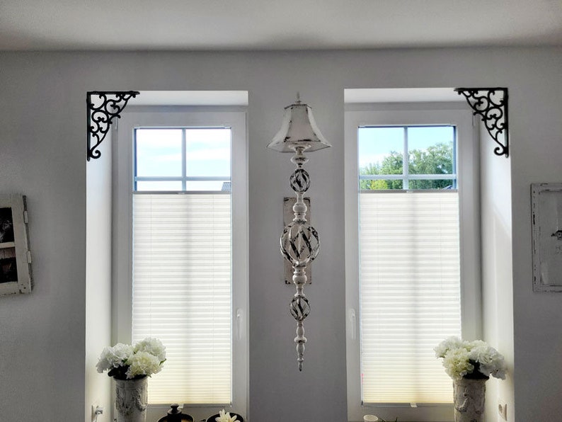 May include: A vertical wall-mounted decorative ornament featuring a distressed white finish with dark accents. The piece consists of a small lampshade-style top, two spherical cage-like sections, and a pointed finial base, positioned between two windows.