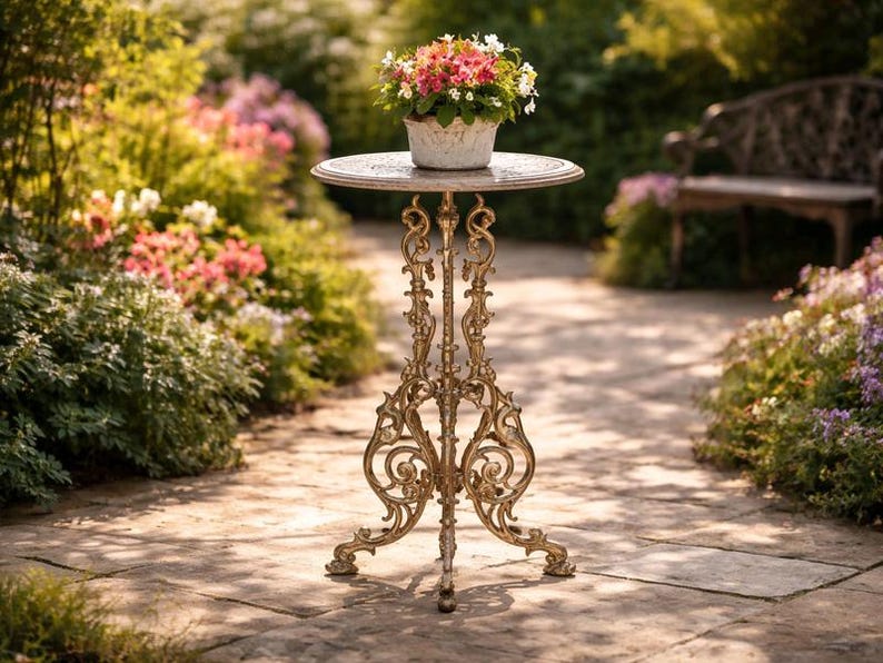 May include: A decorative, ornate, antique-style plant stand with a round top and intricate, golden-colored metalwork. A white pot filled with colorful flowers sits atop the stand, set in a garden setting.