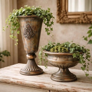 Puede incluir: Dos jardineras de color bronce, de estilo antiguo, con plantas verdes. La jardinera más alta tiene un diseño de flor de lis. La jardinera más corta tiene forma de cuenco. Ambas están sobre una mesa de madera blanca.