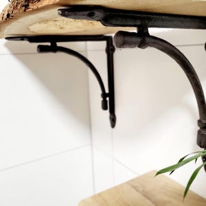 Könnte beinhalten: Zwei schwarze Metall-Regalhalterungen mit gebogenem Design. Die Halterungen sind an einem Holzregal befestigt.