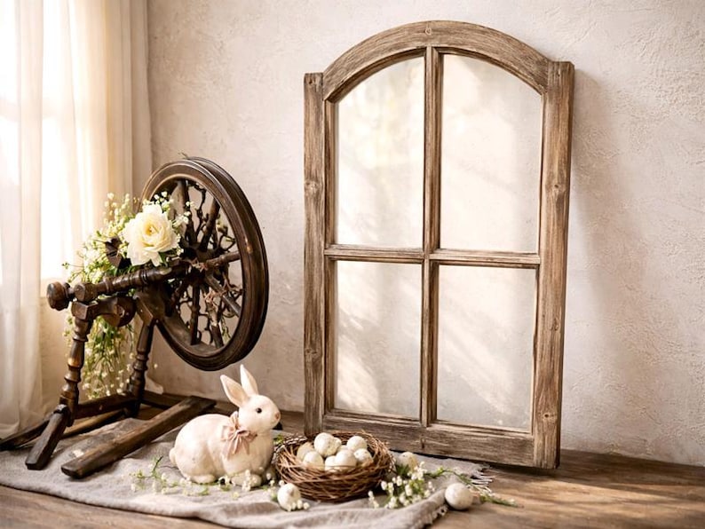 May include: A rustic wooden spinning wheel adorned with white roses and greenery, alongside a white bunny with a pink bow, a nest of eggs, and a weathered wooden window frame. The scene evokes a vintage, farmhouse aesthetic.