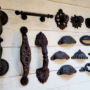 May include: Collection of antique-style, dark brown cast iron hardware. Includes door knockers, handles, knobs, and pulls. The hardware features ornate designs and various shapes, suitable for furniture or doors.