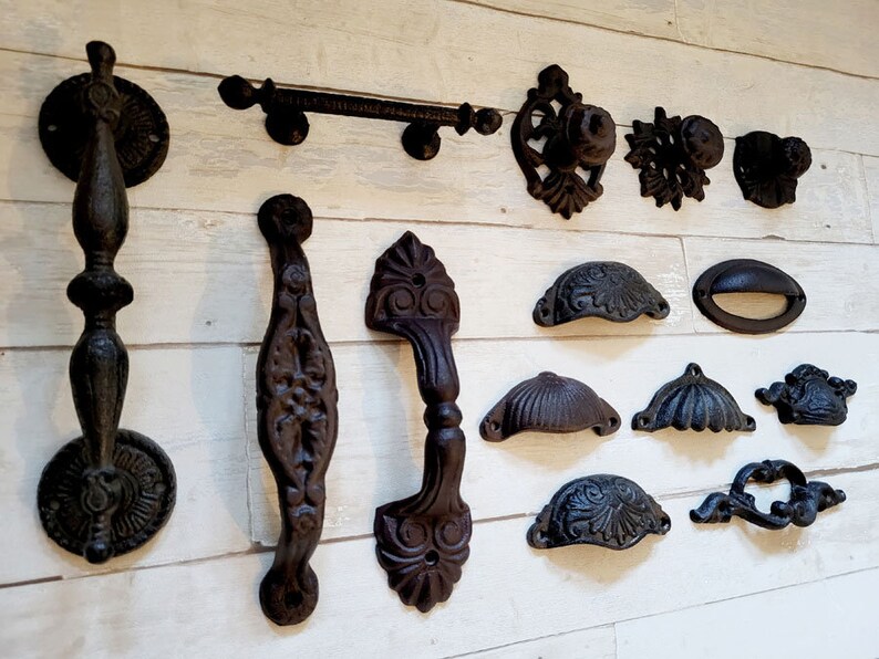 May include: Collection of antique-style, dark brown cast iron hardware. Includes door knockers, handles, knobs, and pulls. The hardware features ornate designs and various shapes, suitable for furniture or doors.