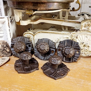 Könnte beinhalten: Ein Set aus vier dunkelbraunen gusseisernen Schubladenknöpfen, jeweils mit einem strukturierten, runden Knopfkopf auf einer quadratischen Rückplatte mit dekorativen Blumenprägungen und zwei vorgebohrten Schraublöchern.