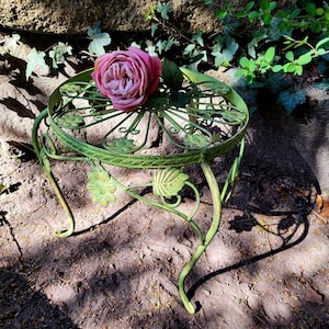 Puede incluir: Un soporte de metal verde para plantas con un diseño floral decorativo, que sostiene una gran rosa rosa. El soporte tiene patas curvas y está colocado sobre una superficie de tierra, con vegetación en el fondo.