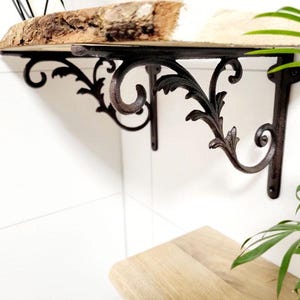 Könnte beinhalten: Zwei schwarze Metall-Regalhalterungen mit verzierten Schnörkeldesigns. Die Halterungen sind an einem Holzregal befestigt.