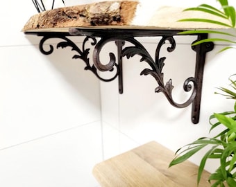 May include: A dark brown metal shelf bracket featuring an ornate scrollwork design with leaf-like details. The bracket supports a thick wooden shelf with a natural, rough-hewn live edge. The metal structure is mounted to a white wall surface.