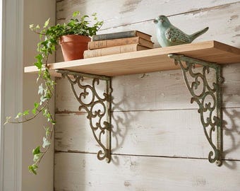 Puede incluir: Estantería de madera con soportes metálicos ornamentados. Exhibe una planta en maceta, libros y una figura de pájaro azul. Los soportes tienen un aspecto envejecido, y la estantería está fijada a una pared de madera pintada de blanco. La estantería mide unos 76 cm.