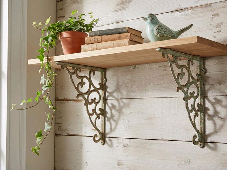 May include: A light wood wall shelf supported by two ornate, weathered green metal brackets with scrollwork designs. On the shelf sits a terracotta pot with a trailing green ivy plant, a stack of three vintage books, and a small ceramic bird figurine.