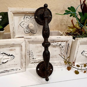 May include: A dark brown, ornate metal door handle with a decorative design. The handle has a long, vertical grip and circular mounting plates. The background includes white wooden frames with floral embellishments, suggesting a rustic or vintage aesthetic.