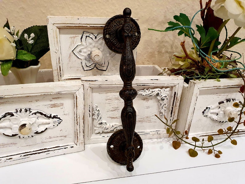 May include: A dark brown, ornate metal door handle with a decorative design. The handle has a long, vertical grip and circular mounting plates. The background includes white wooden frames with floral embellishments, suggesting a rustic or vintage aesthetic.