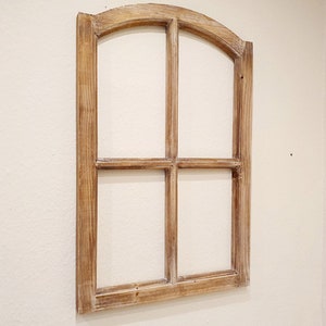 May include: A decorative wooden window frame with a light brown finish. The frame has an arched top and is divided into four panes. It is mounted on a cream-colored wall, adding a rustic touch to the decor.