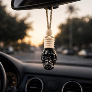 May include: A black skull-shaped glass car air freshener bottle hanging from a rearview mirror by a tan braided cord. The bottle features a light-coloured wooden cap and a small wooden bead, suspended above a car dashboard in a blurred outdoor setting.