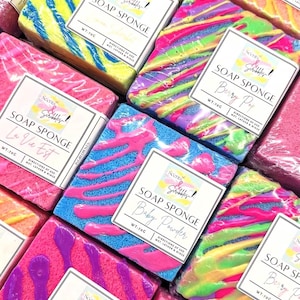 May include: A selection of vibrant soap sponges in shades of pink, blue, yellow, and green. Each sponge is individually wrapped and labelled with the "Scents of Scribbles" brand, with scents such as "Berry Pop" and "Baby Powder". Each sponge weighs 70g.
