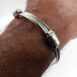 Può includere: Un braccialetto color argento con un design a più fili. Il braccialetto presenta una serie di fili paralleli tenuti insieme da elementi terminali decorativi e avvolti. Il gioiello è mostrato sul braccio di una persona.