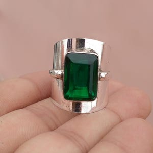 Puede incluir: Un anillo ancho de tono plateado que presenta una gran piedra preciosa rectangular de color verde esmeralda facetada en el centro. La banda tiene un acabado metálico liso y pulido con un engaste vertical prominente que sujeta la piedra.