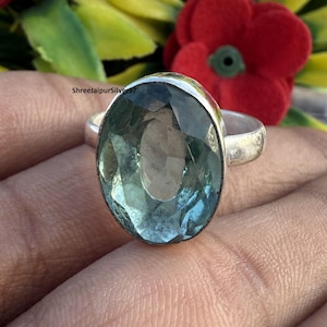 Może przedstawiać: Srebrny pierścionek z dużym, owalnym, jasnoniebieskim kamieniem szlachetnym. Kamień szlachetny ma wiele faset, które odbijają światło. Pierścionek ma prosty, polerowany srebrny pas. Tło jest rozmyte, zielone i czerwone.